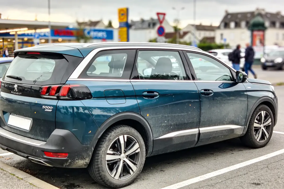 SUV Peugeot 5008 bleu garé sur un parking, magasins en arrière-plan, ciel nuageux.