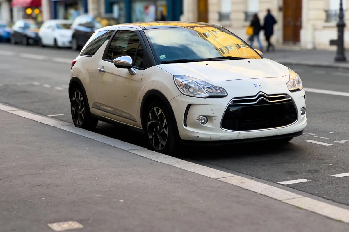 Voiture blanche citadine garée en bordure de trottoir, rue urbaine avec immeubles en arrière-plan.