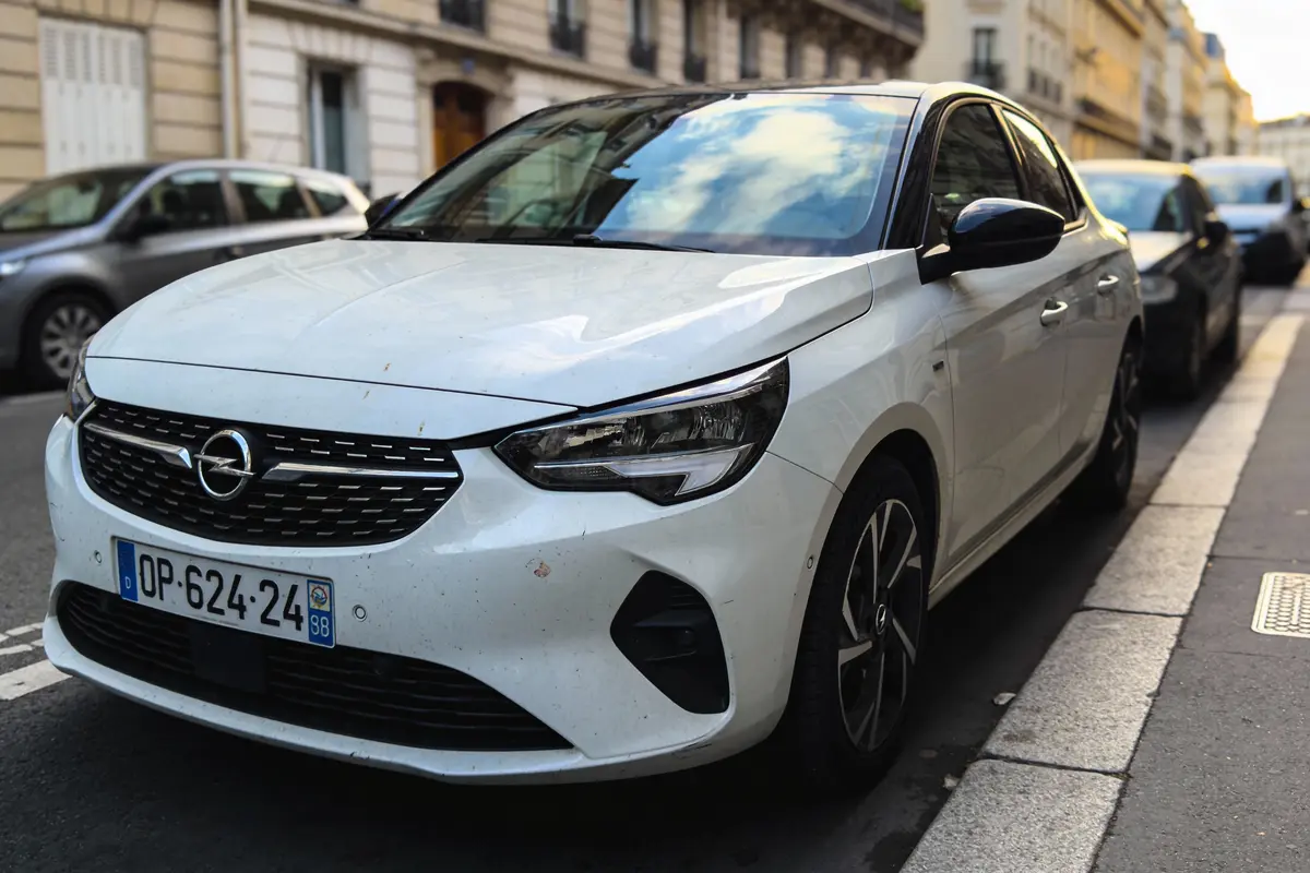 Voiture Opel blanche garée dans une rue urbaine, bâtiments résidentiels en arrière-plan, plaques d'immatriculation visibles.
