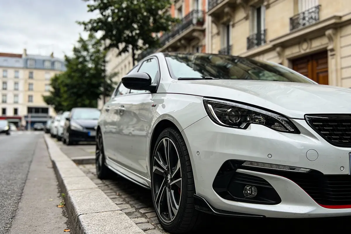 Voiture blanche moderne garée dans une rue urbaine avec bâtiments et arbres en arrière-plan.