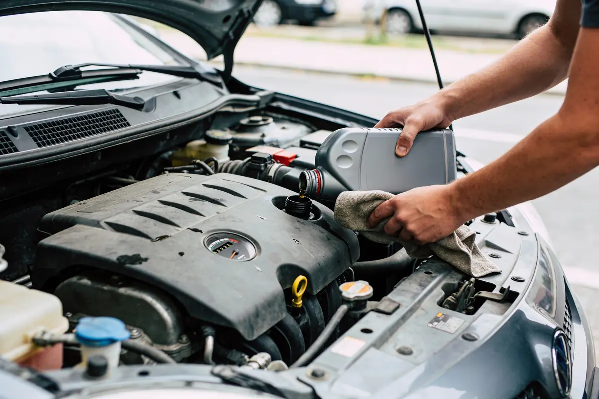 Personne versant de l'huile moteur dans une voiture, capot ouvert, rue en arrière-plan.
