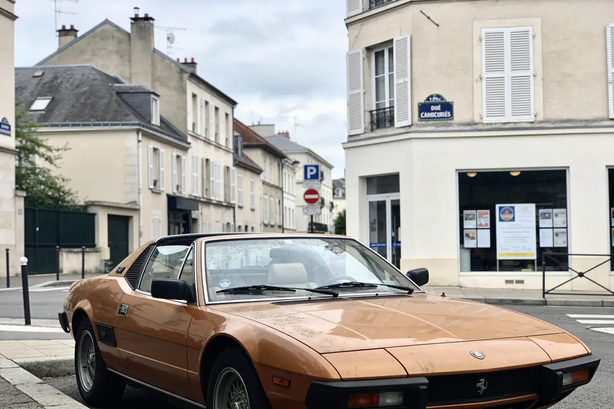 Voiture classique brune stationnée dans une rue calme avec bâtiments résidentiels derrière. Feux et panneaux visibles.