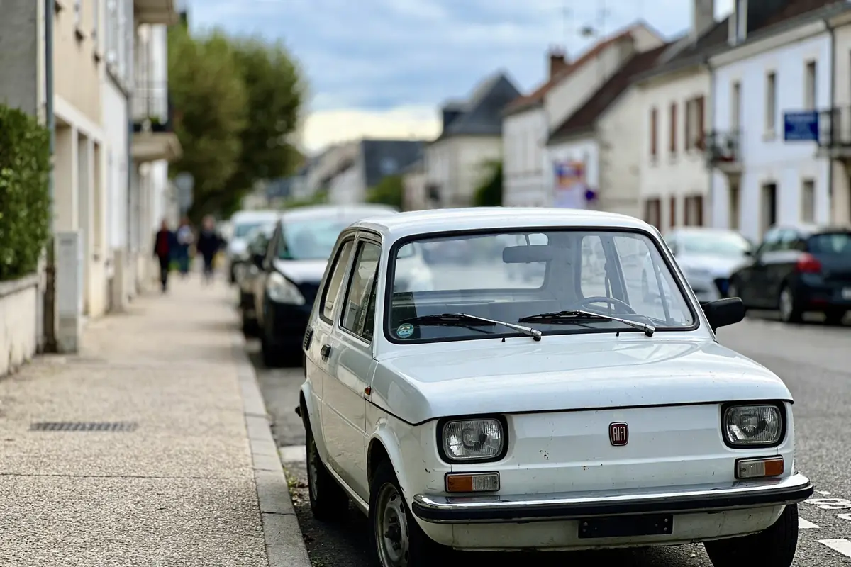 Voiture blanche garée sur trottoir, rue bordée d'arbres, bâtiments résidentiels en arrière-plan. Ciel légèrement nuageux.