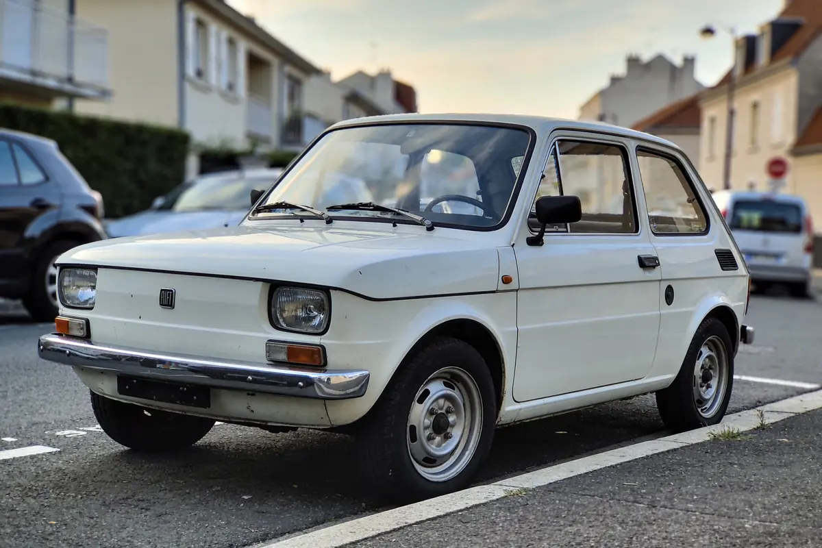 Petite voiture blanche ancienne garée dans une rue résidentielle, ciel dégagé en arrière-plan.