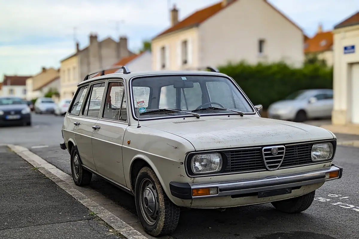 Voiture ancienne stationnée dans une rue résidentielle, maisons en arrière-plan, ciel légèrement nuageux.