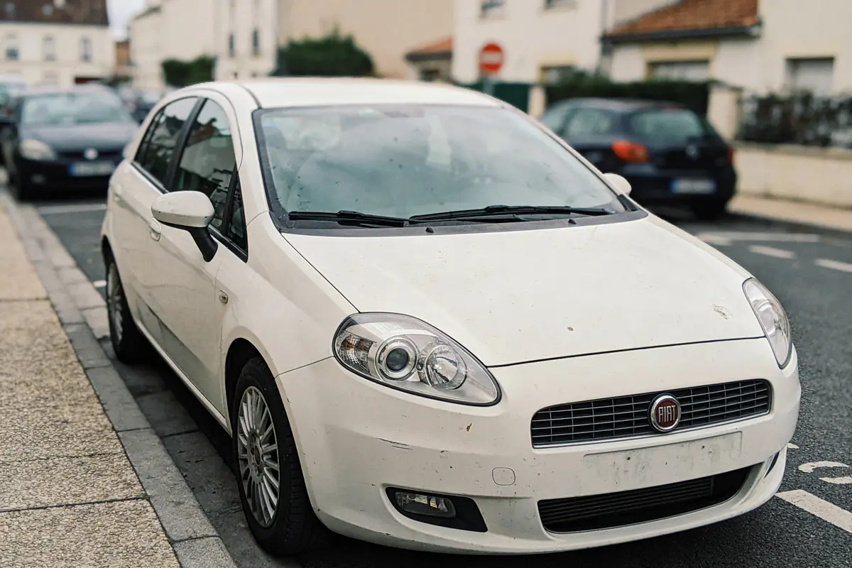 Voiture blanche Fiat garée sur la rue résidentielle, entourée d'autres voitures et bâtiments en arrière-plan.