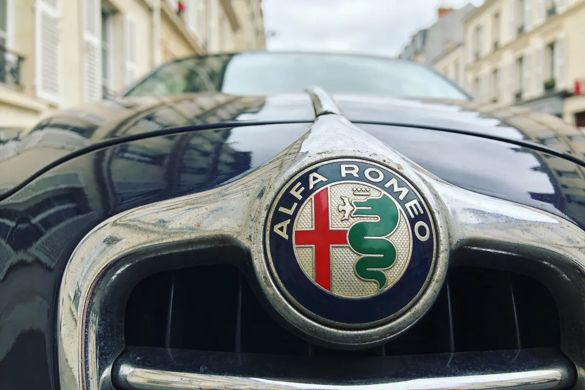 Gros plan sur le logo Alfa Romeo sur le capot d'une voiture, rue résidentielle en arrière-plan.