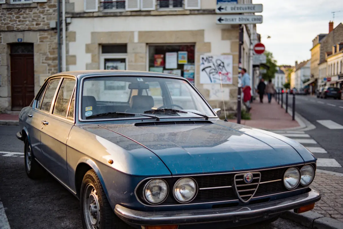 Voiture ancienne bleue garée dans une rue urbaine avec bâtiments et panneaux de signalisation en arrière-plan.