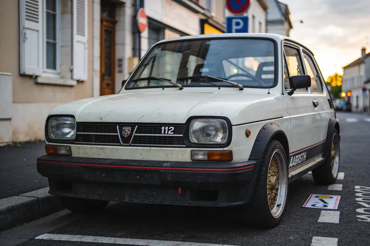 Voiture rétro blanche Abarth 112 garée en ville, au bord d'une rue pavée, à côté de bâtiments.