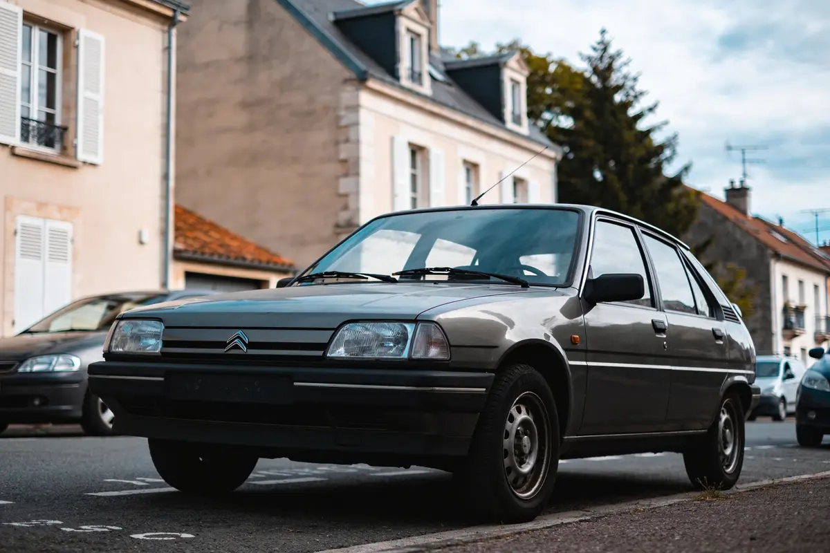 Voiture Citroën grise stationnée devant maisons anciennes, rue urbaine calme, ciel partiellement nuageux.