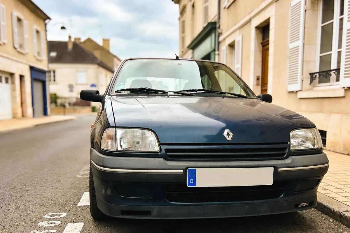 Vieille Renault grise garée dans une rue calme, bâtiments en arrière-plan, ciel nuageux.
