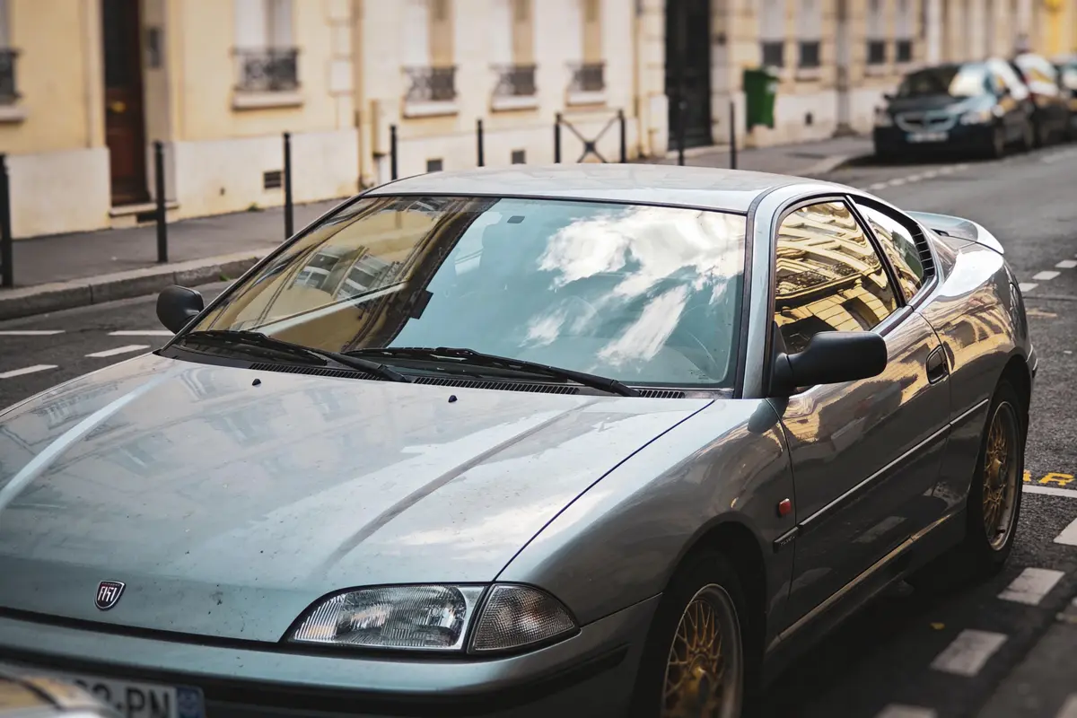Voiture grise stationnée sur rue urbaine, bâtiments reflétés dans pare-brise, ciel nuageux visible.