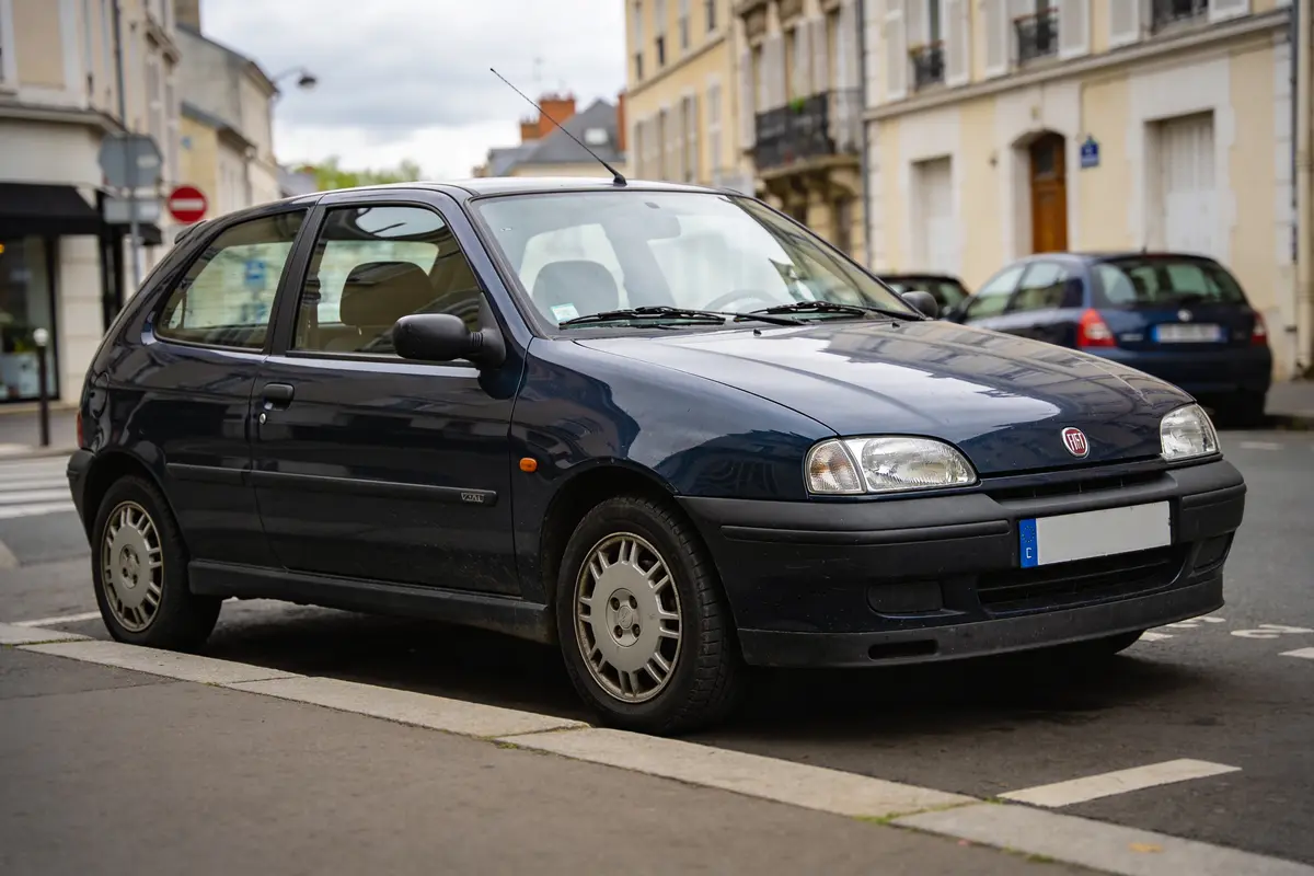 Petite voiture noire stationnée dans une rue urbaine, bâtiments résidentiels en arrière-plan, temps nuageux.