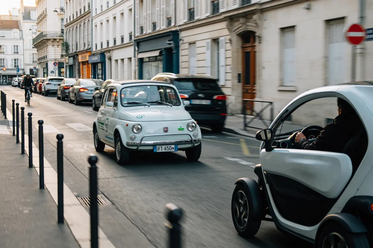 Deux petites voitures blanches et un cycliste circulent dans une rue urbaine bordée d'immeubles.