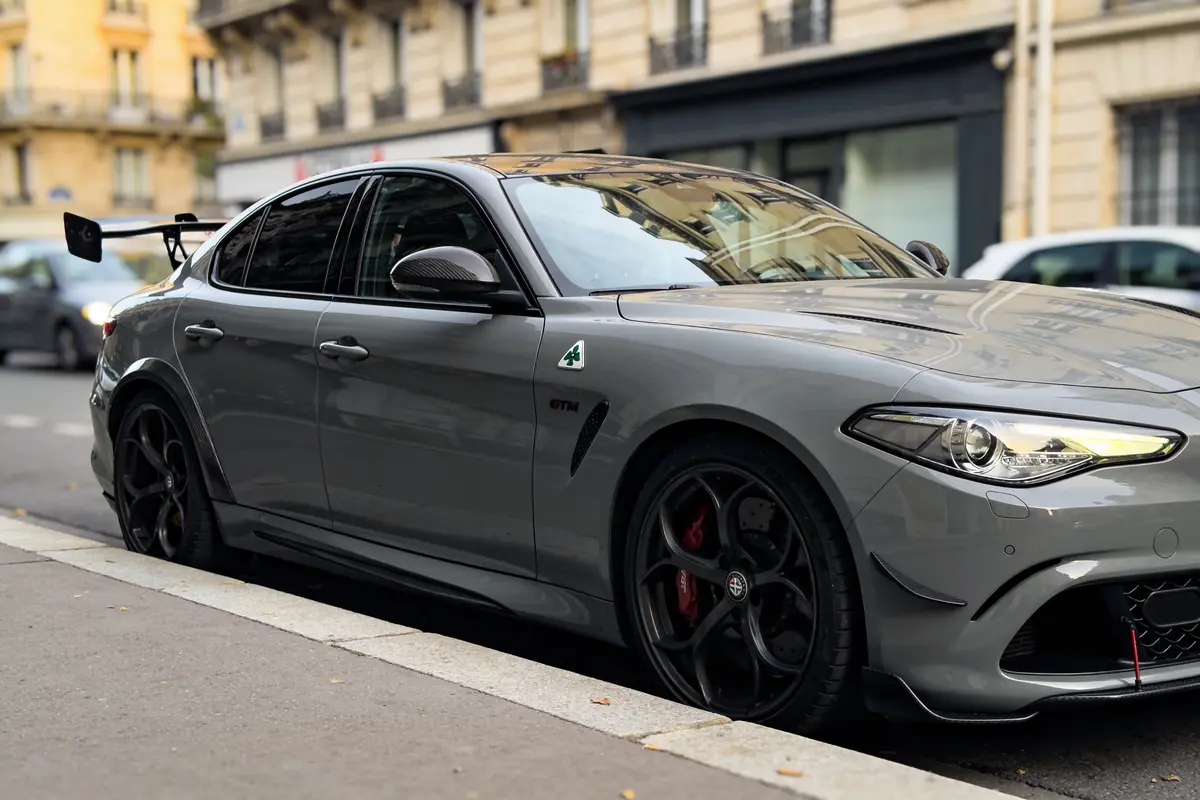 Voiture grise sportive avec aileron, garée en rue urbaine, fond de bâtiments et voitures.