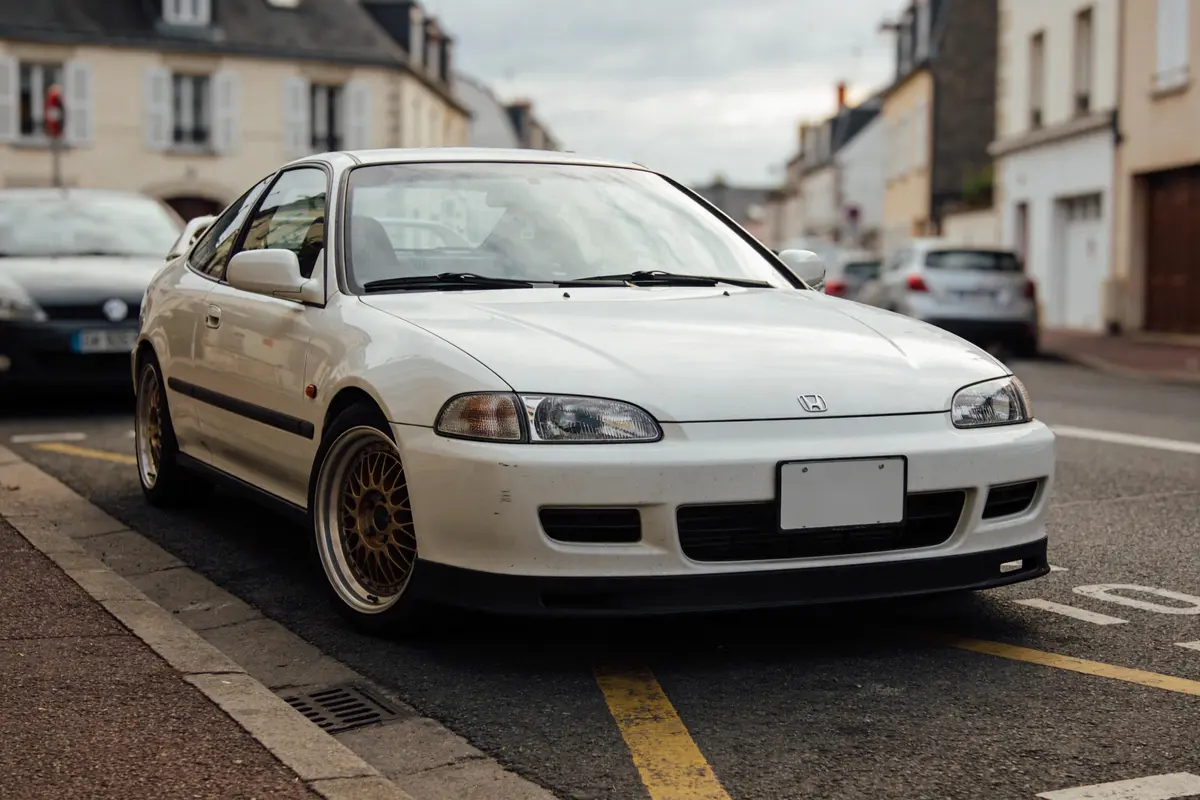 Voiture blanche Honda garée en ville, avec jantes dorées et bâtiments en arrière-plan.