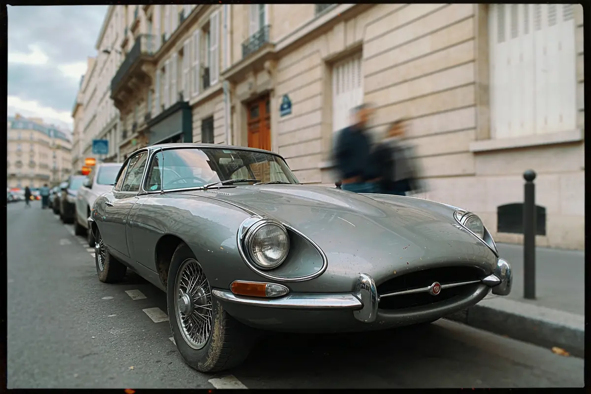Voiture classique grise garée dans une rue parisienne, avec bâtiments et passants en arrière-plan flouté.