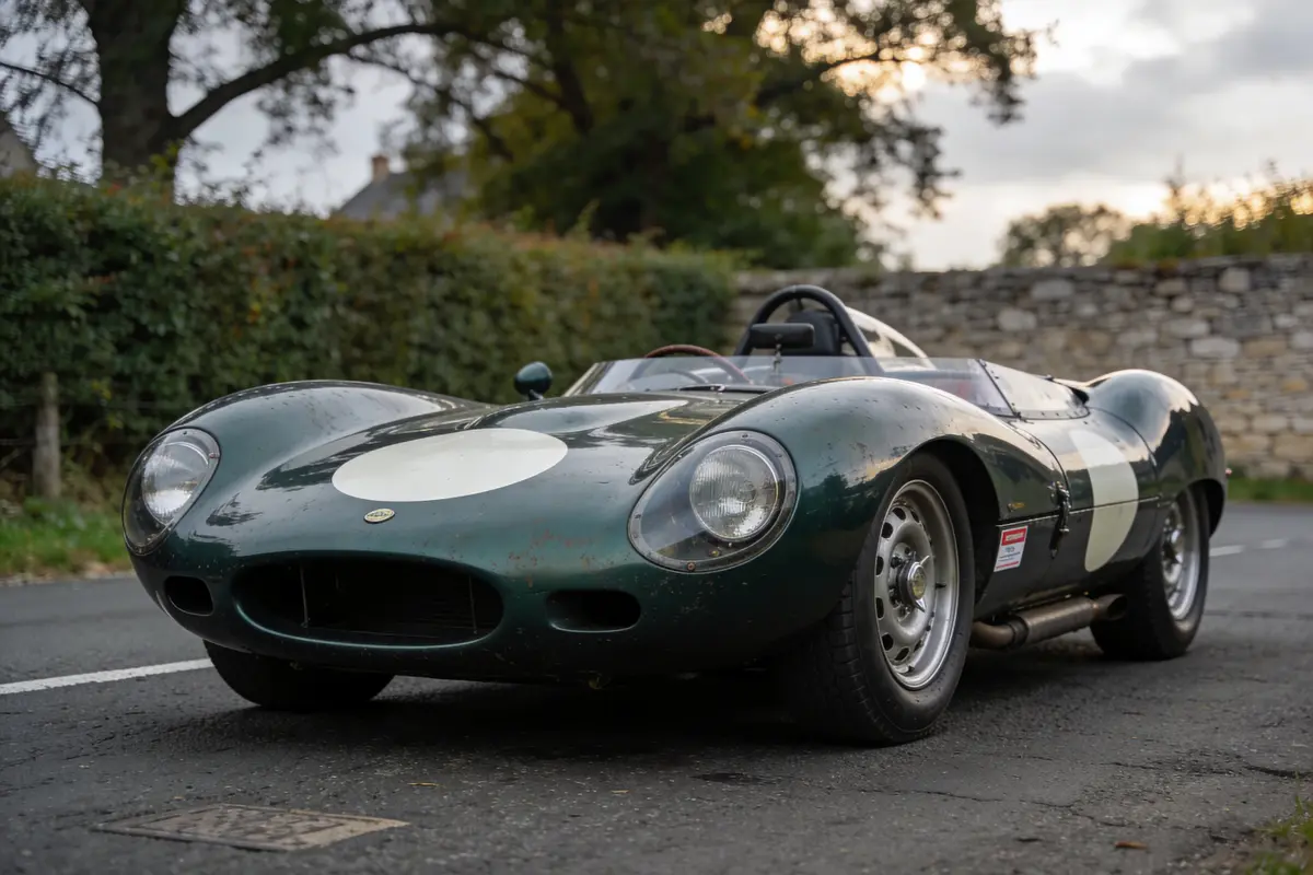 Voiture de course classique verte, garée sur une route, devant un mur en pierre et des arbres.
