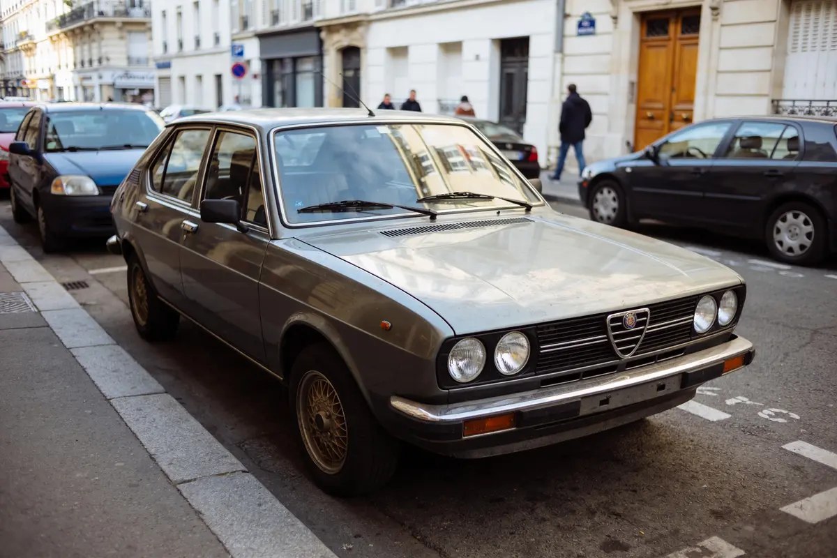 Voiture ancienne grise garée dans une rue urbaine, bâtiments et passants en arrière-plan.