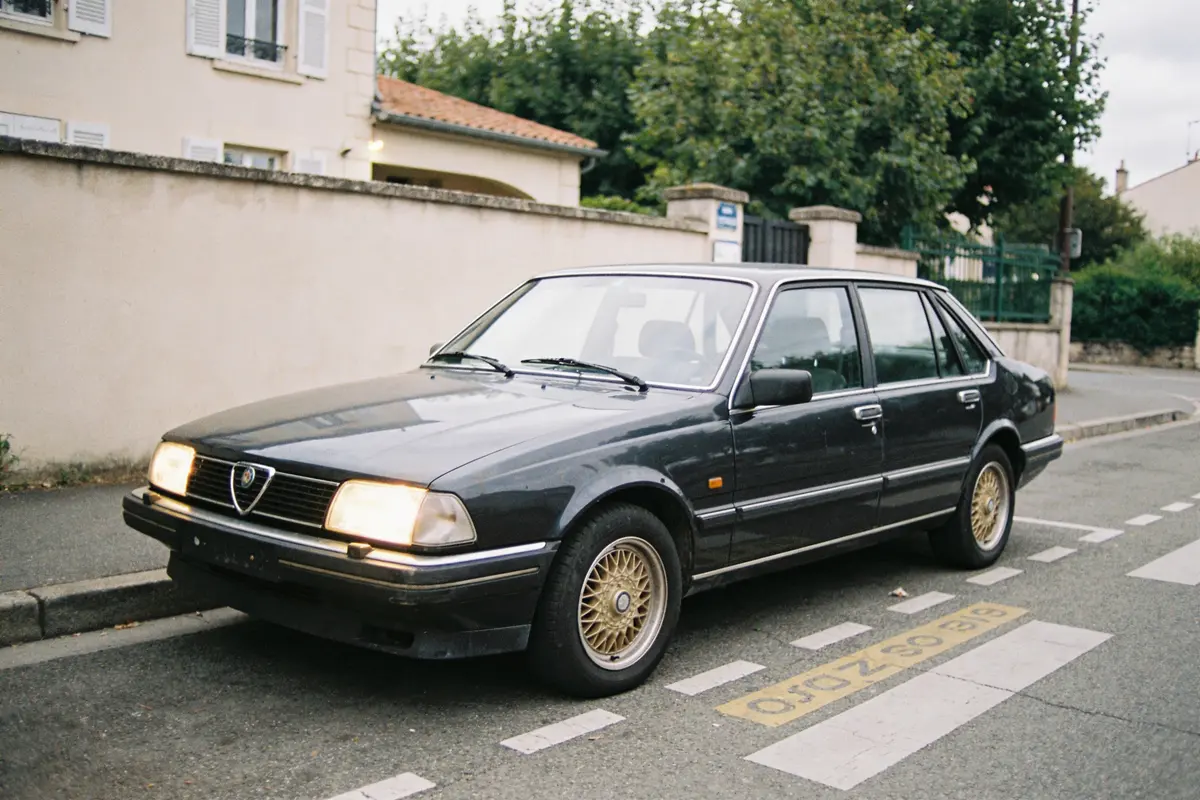Voiture ancienne noire garée sur la route devant une maison, phares allumés.
