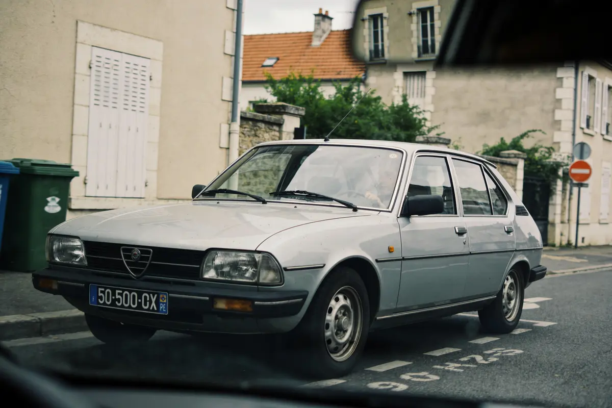 Voiture ancienne grise stationnée dans une rue, bâtiments et poubelles visibles en arrière-plan.