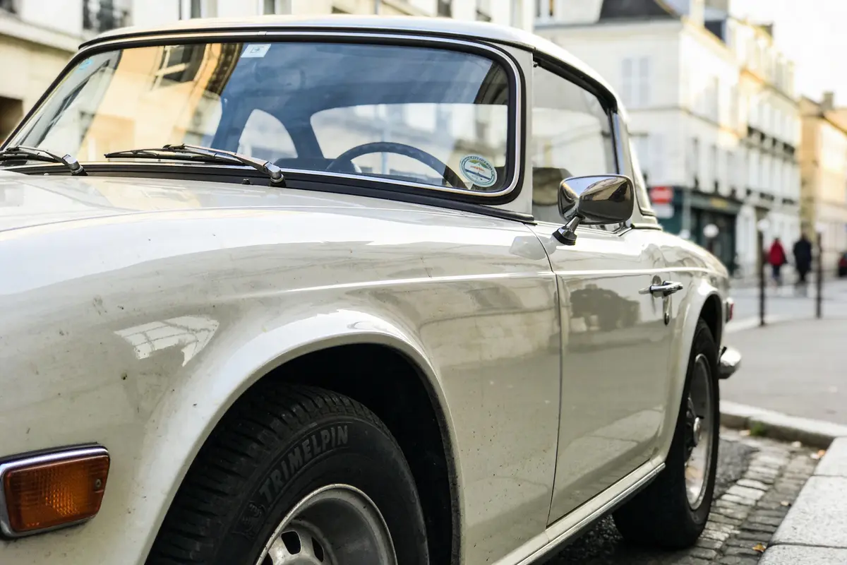 Voiture classique blanche garée sur la rue pavée en ville, bâtiments flous en arrière-plan.