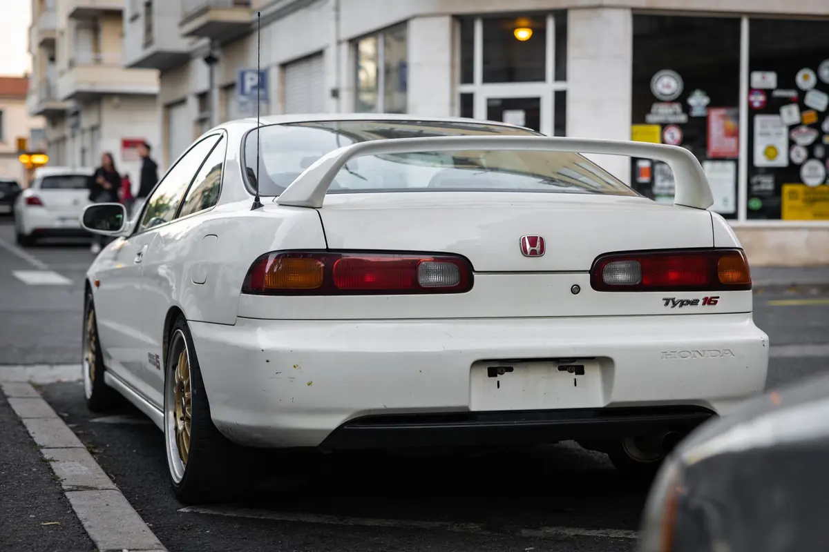 Voiture blanche, Honda, stationnée en ville, aileron arrière visible, bâtiment en arrière-plan, route pavée, autocollants.