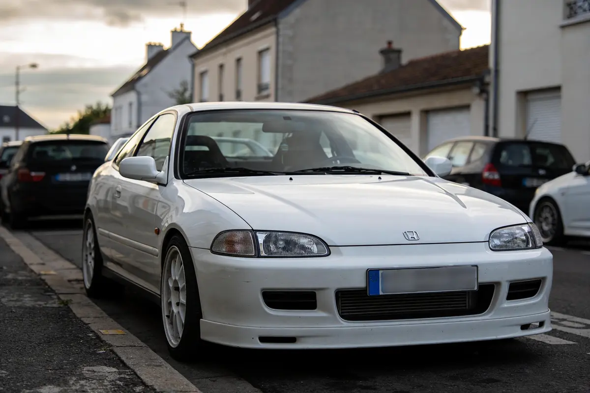 Voiture blanche garée dans une rue résidentielle, maisons en arrière-plan.