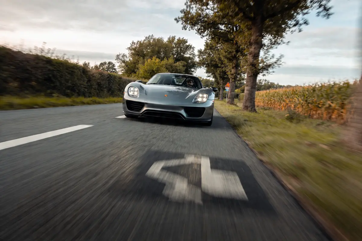 Voiture de sport grise sur route campagnarde, bordée d'arbres et de champs, sous ciel nuageux.