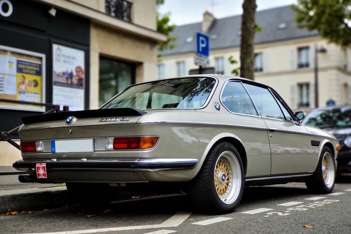 Voiture classique BMW 3.0 CSL grise, stationnée dans la rue devant des bâtiments résidentiels.