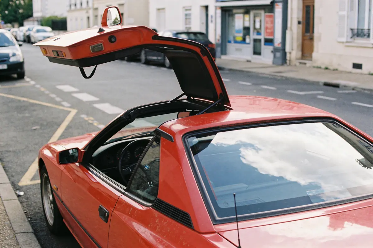 Voiture rouge avec portière type papillon sur rue, reflet de nuages sur pare-brise.