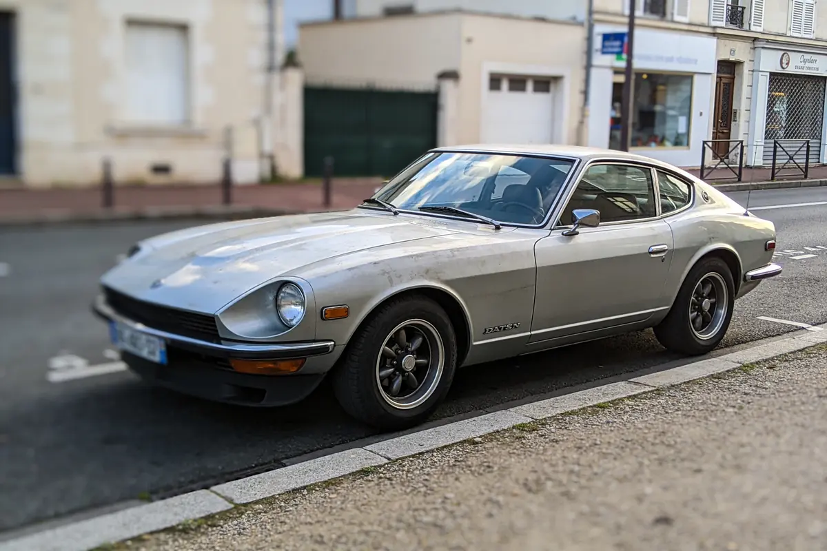 Voiture ancienne Datsun grise garée dans une rue résidentielle, bâtiments en arrière-plan flou.