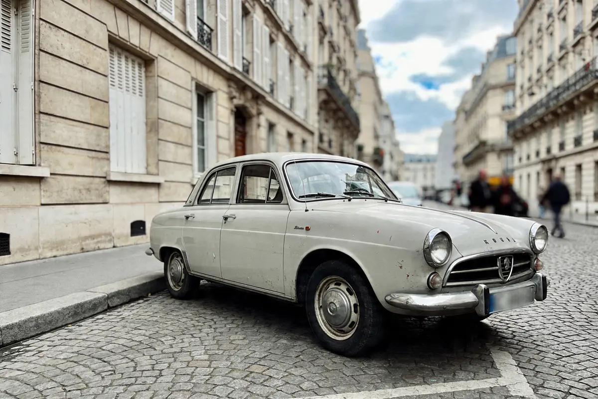 Voiture ancienne blanche garée sur rue pavée parisienne, immeubles haussmanniens en arrière-plan, ciel nuageux.