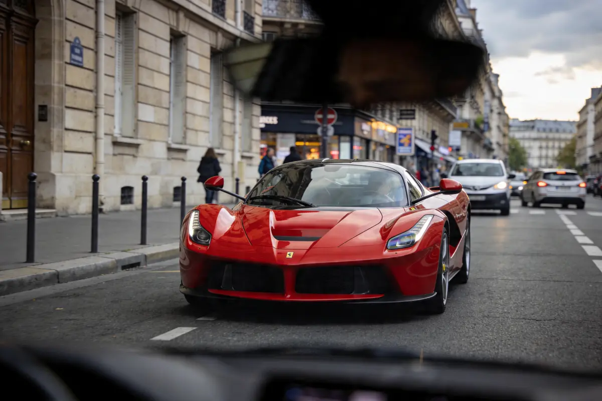 Voiture de sport rouge circulant dans une rue parisienne, avec bâtiments et quelques piétons en arrière-plan.