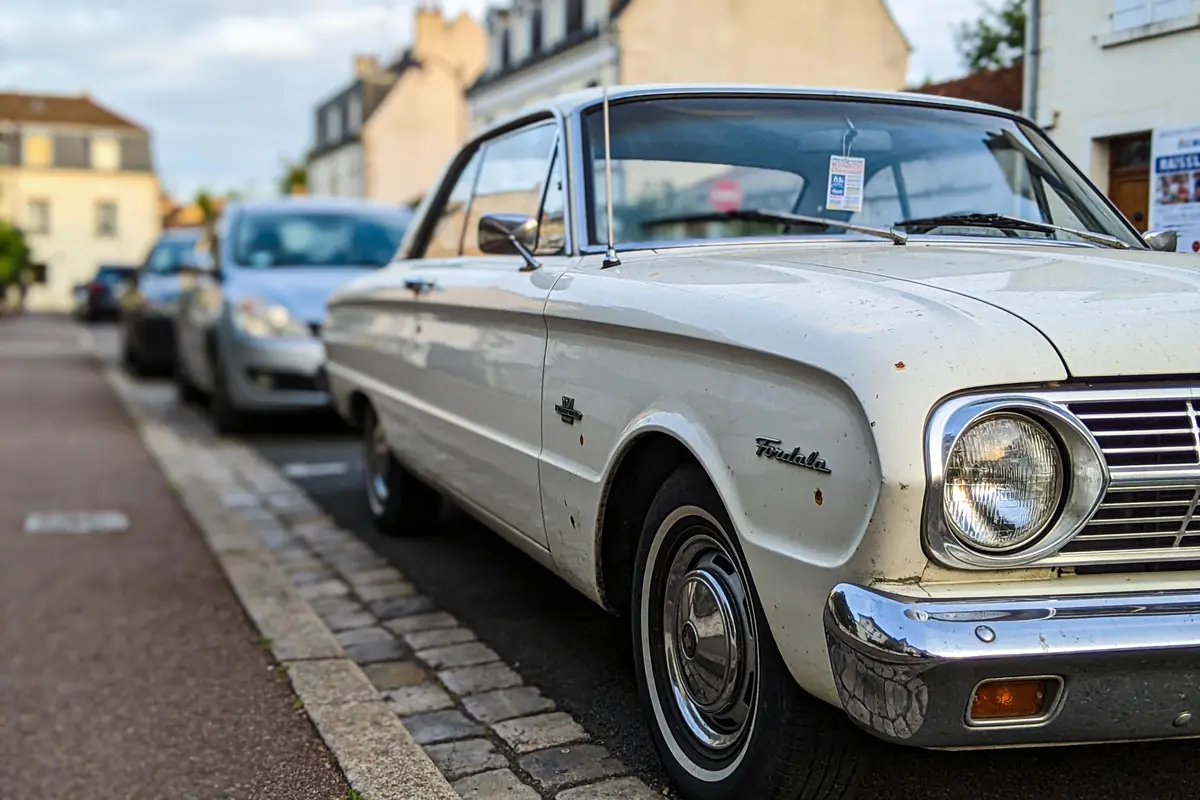 Vieille voiture blanche garée en ville, avec bâtiments résidentiels flous en arrière-plan.