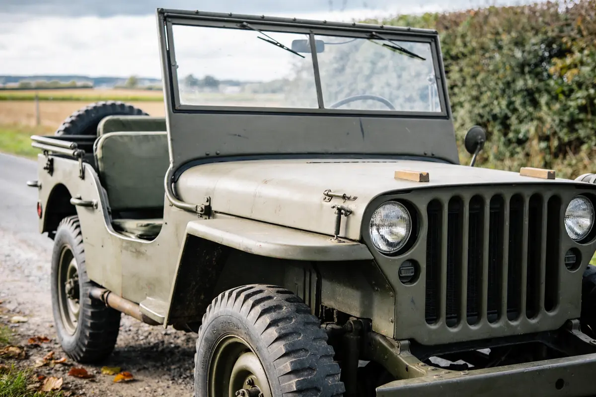 Jeep militaire ancienne, couleur kaki, stationnée sur route de campagne bordée de végétation.