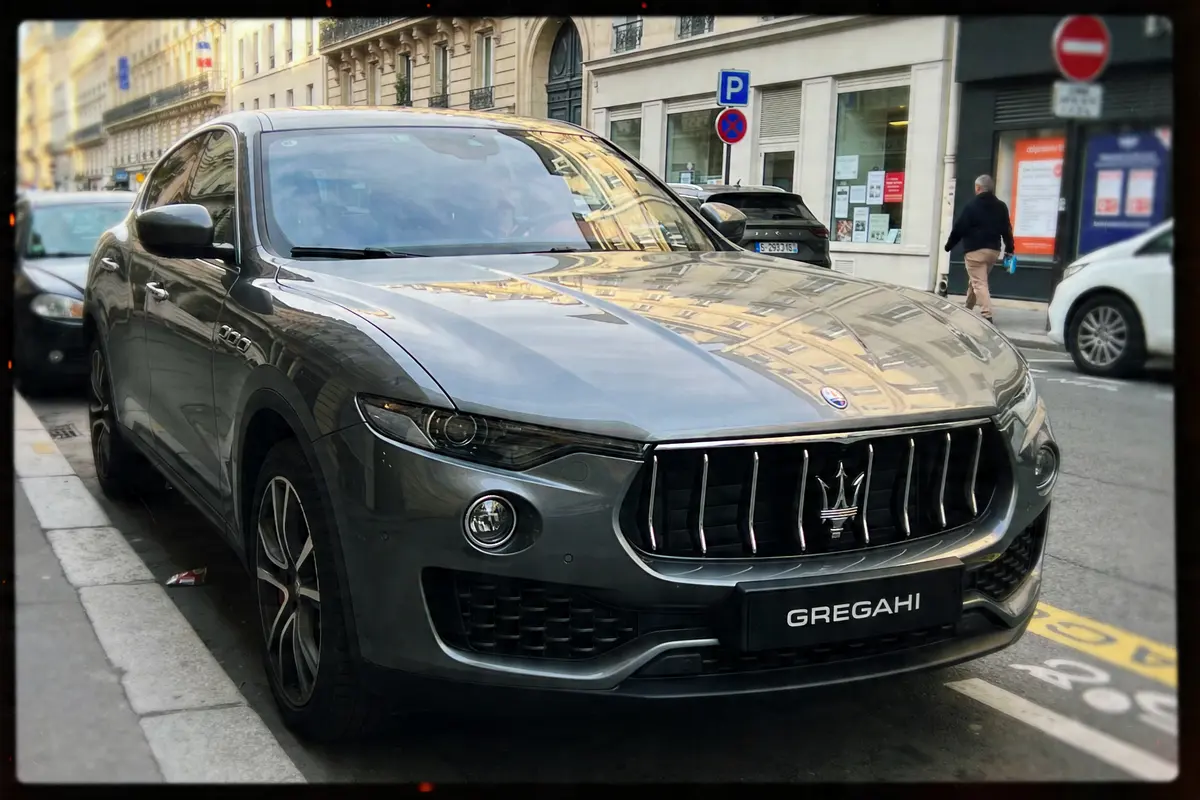 Voiture grise Maserati stationnée en ville, devant un bâtiment avec pancartes et personnes.