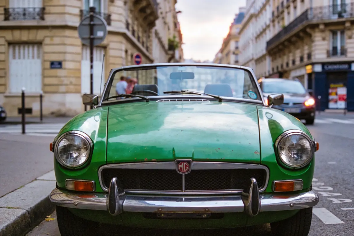 Voiture de collection verte, décapotable, stationnée dans une rue urbaine avec bâtiments historiques environnants.