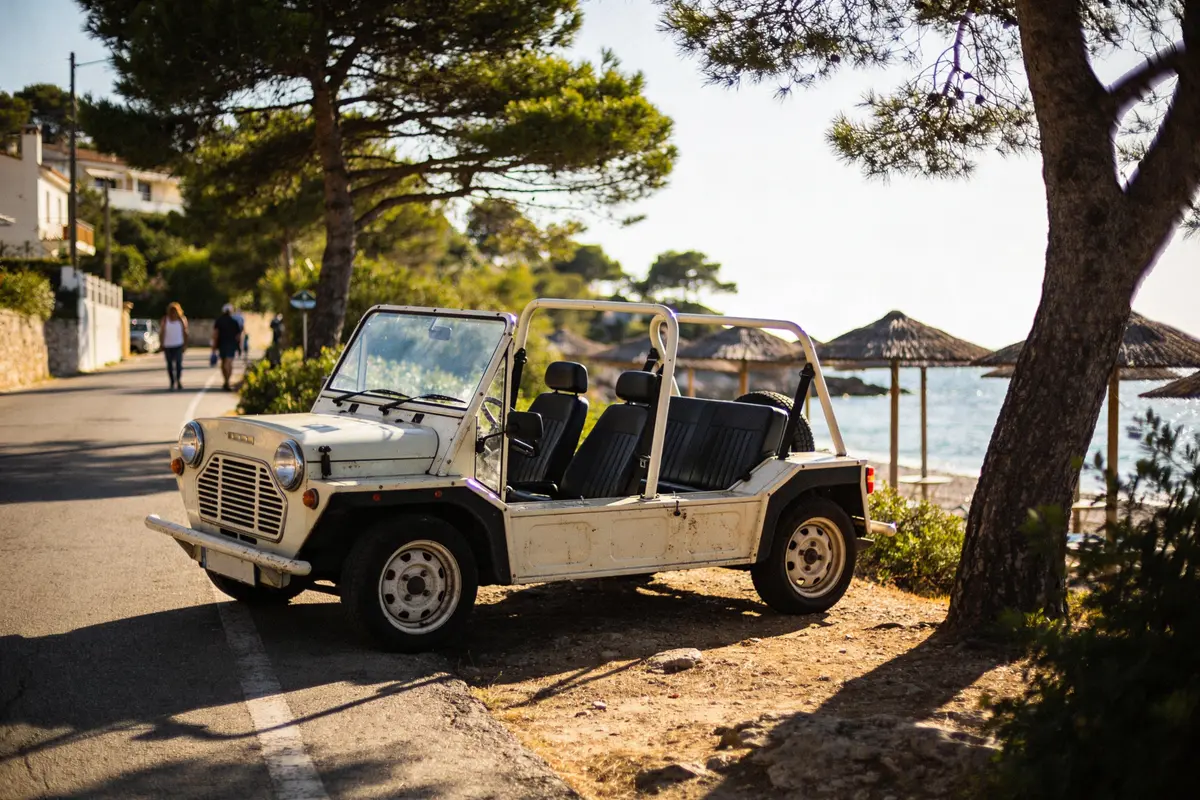 Voiture vintage blanche décapotable garée près de la plage, route bordée d'arbres, parasols en arrière-plan.