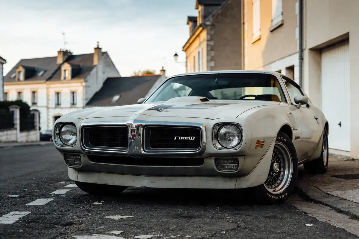 Voiture classique blanche garée dans une rue résidentielle bordée de maisons anciennes.