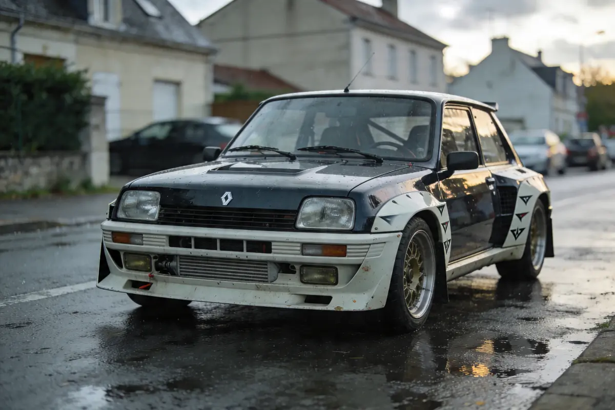 Voiture Renault 5 noire sur route mouillée, bâtiments résidentiels en arrière-plan, ciel nuageux.