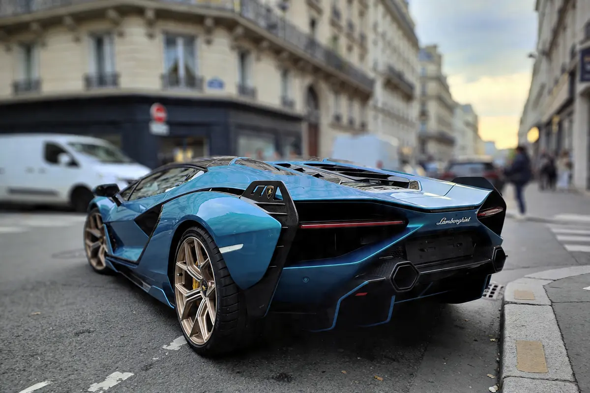 Voiture de sport bleue garée en ville, bâtiment haussmannien en arrière-plan, ciel légèrement couvert.