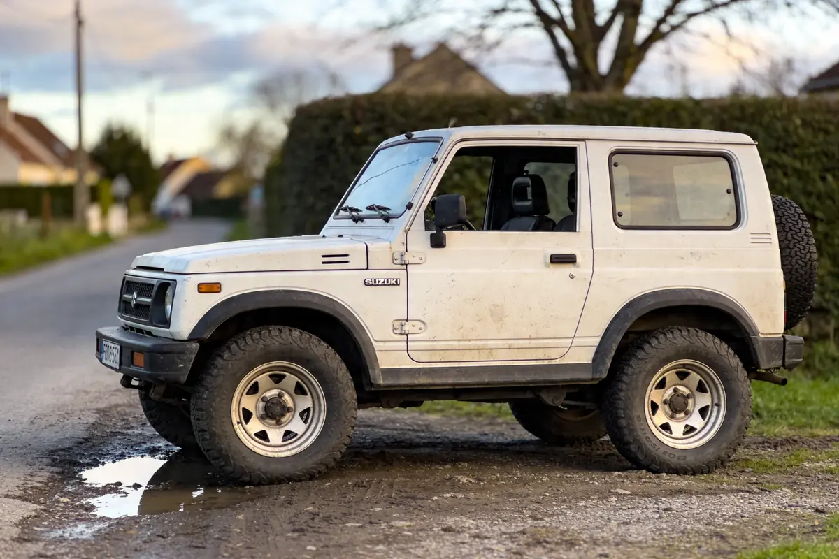 Véhicule tout-terrain blanc garé sur chemin, avec décor de campagne floue en arrière-plan.