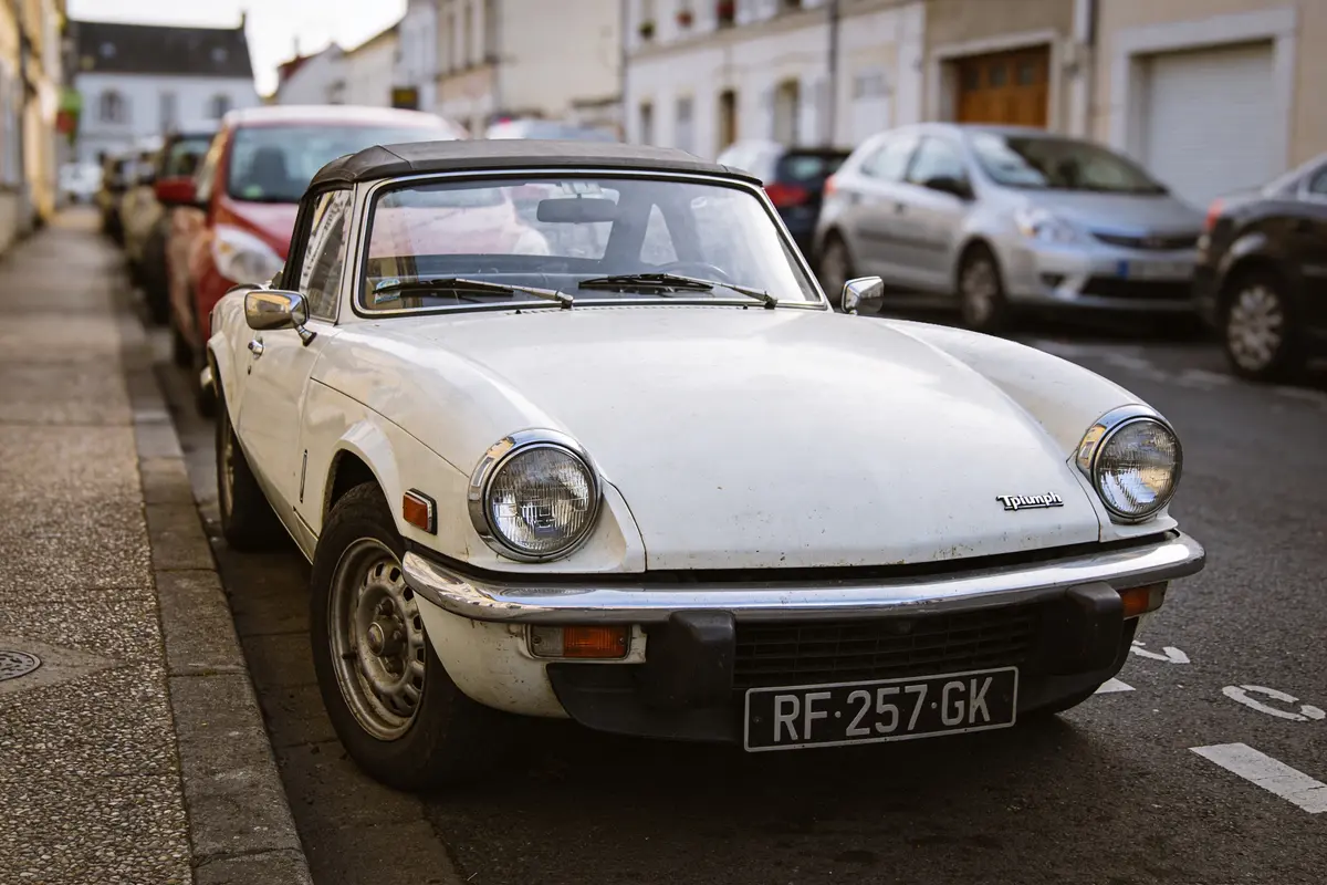 Voiture blanche ancienne, garée en ligne de rue urbaine, bâtiments résidentiels en arrière-plan.