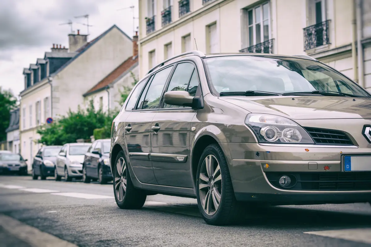 Voiture Renault beige stationnée dans une rue résidentielle avec bâtiments en arrière-plan, ciel nuageux.