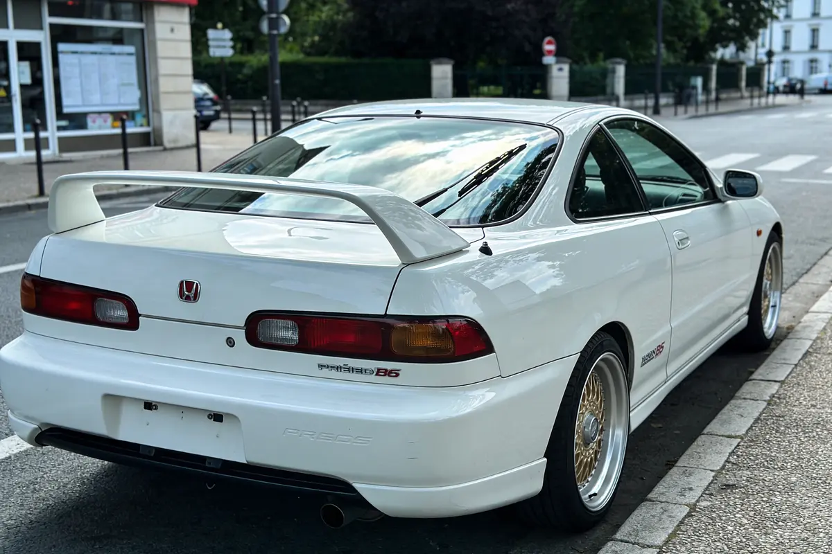 Voiture blanche sportive garée dans la rue, avec aileron arrière et jantes dorées.