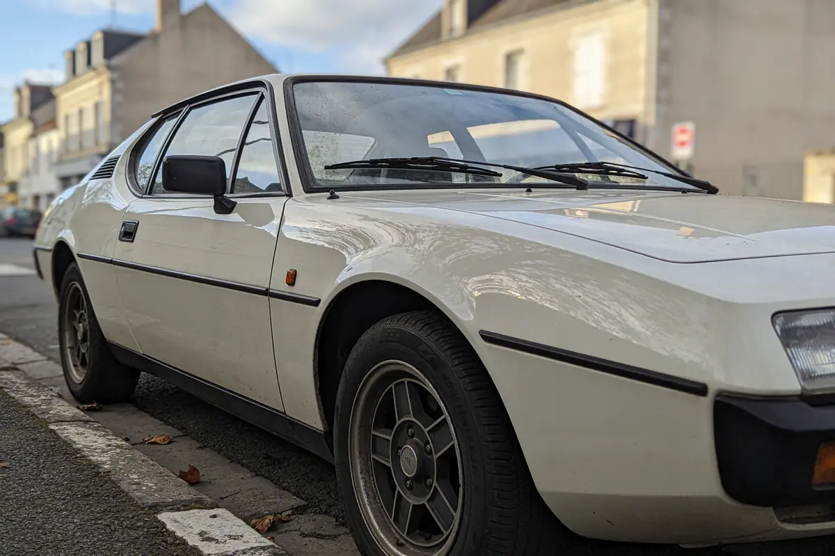 Voiture vintage blanche garée dans une rue résidentielle, bâtiments en arrière-plan.