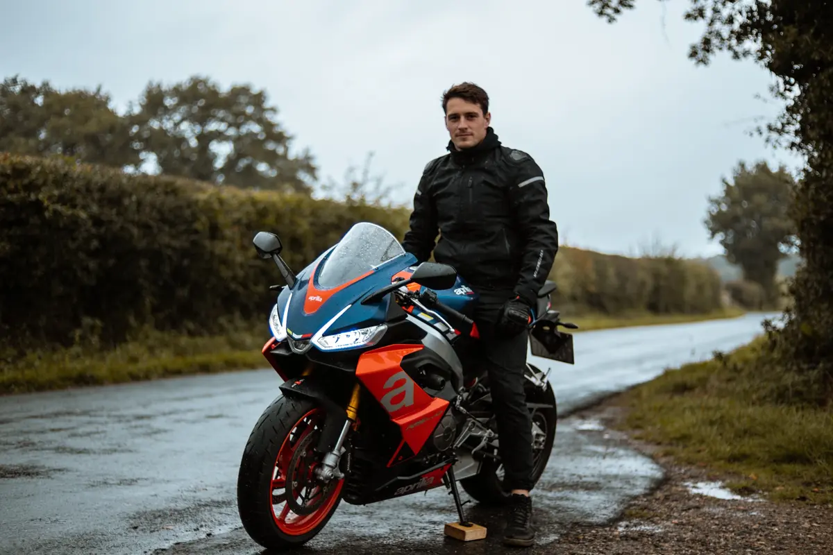 Homme en tenue de moto debout à côté d'une moto sportive colorée sur une route campagnarde.
