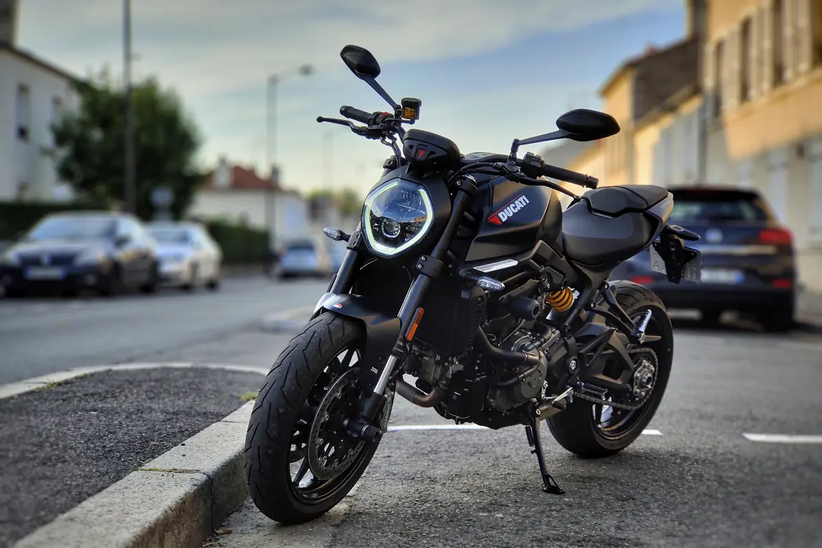 Moto Ducati noire garée sur une route urbaine, avec voitures floues en arrière-plan.