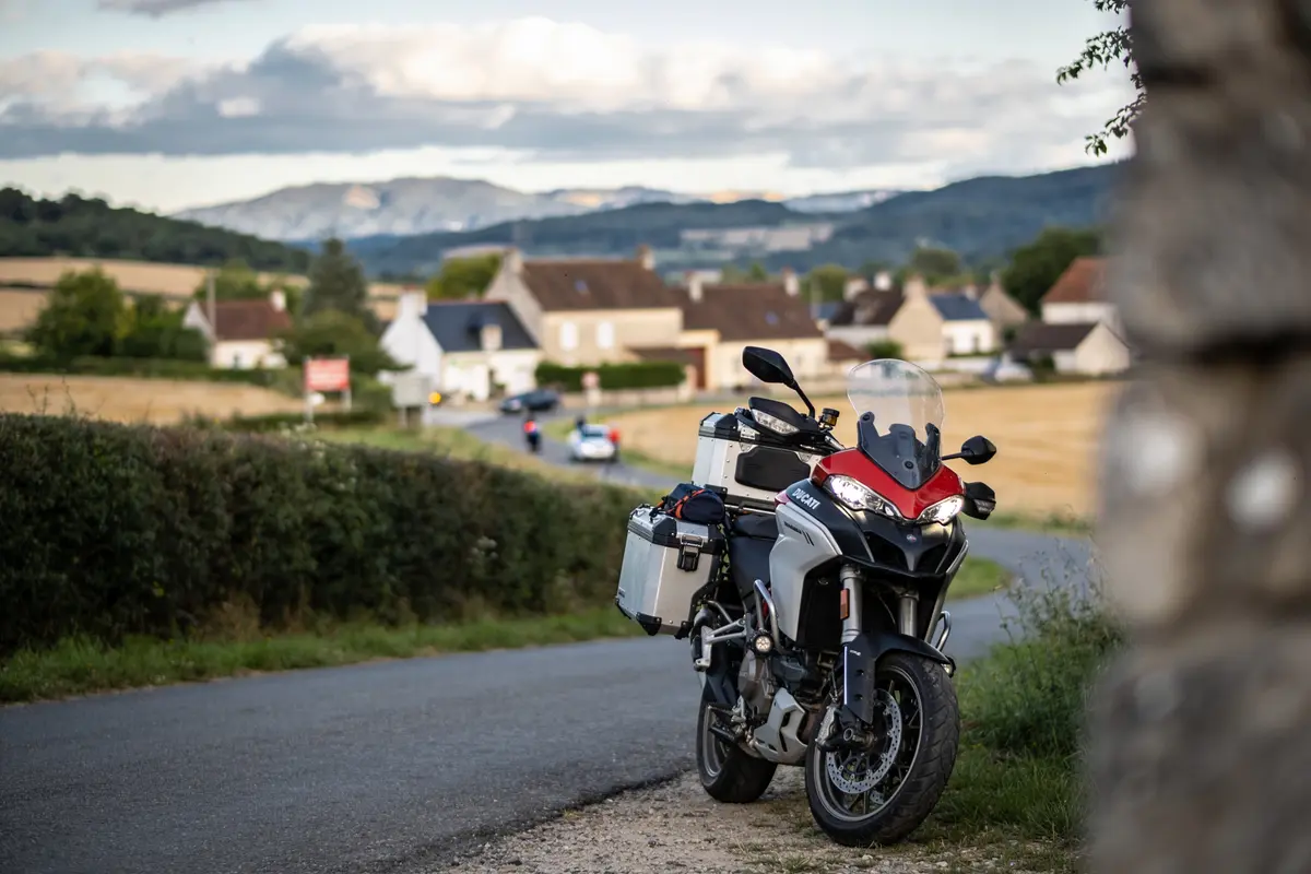 Moto stationnée sur route de campagne, paysage de village et montagnes en arrière-plan. Journée ensoleillée.
